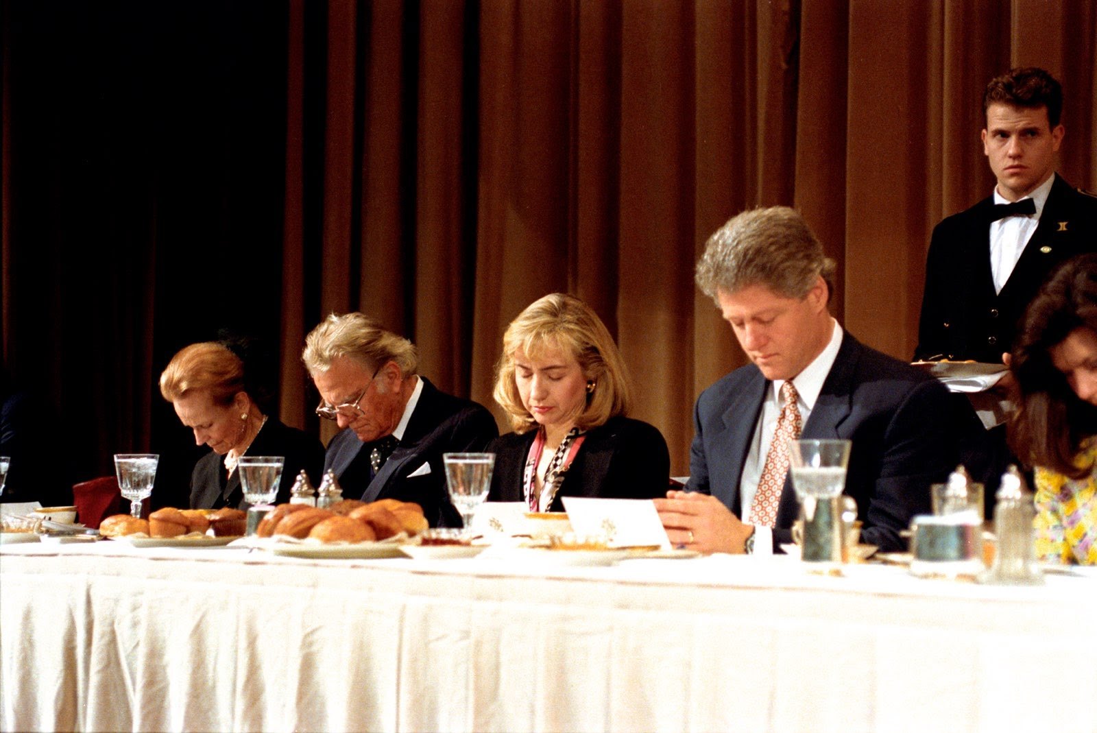 President Bill Clinton and Hillary Clinton with heads bowed in prayer at the Prayer Breakfast